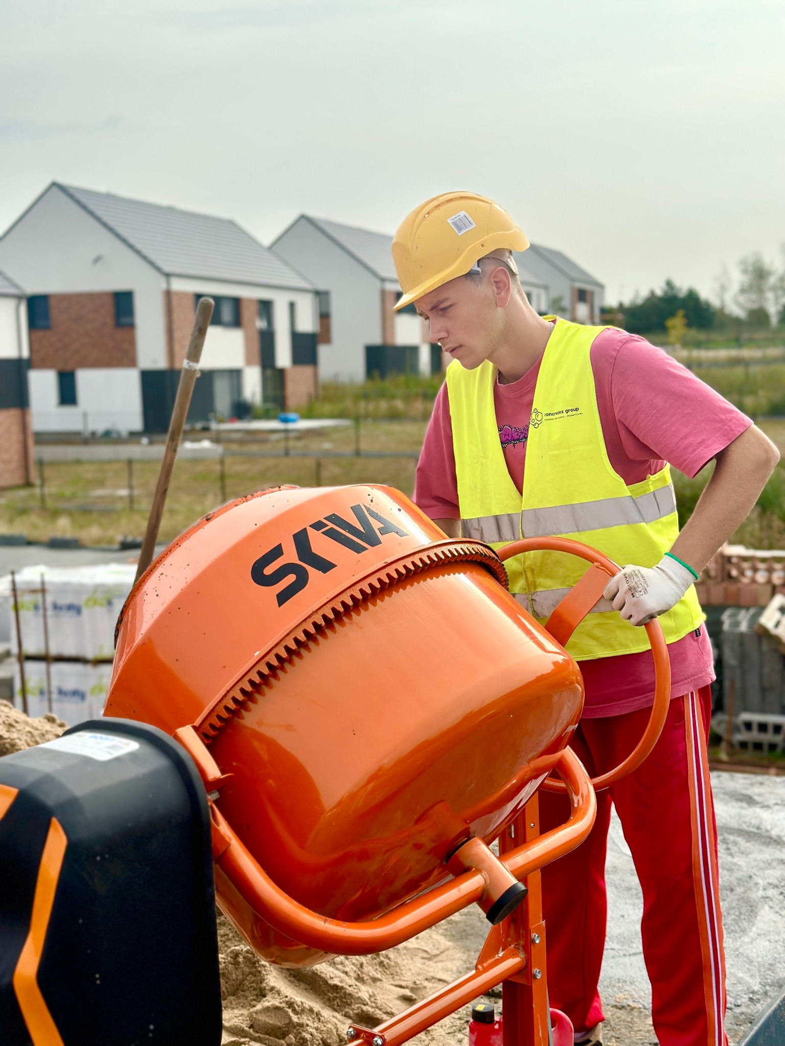 Młody budowlaniec w kasku i kamizelce ochronnej obsługuje pomarańczową betoniarkę marki SKYVA na placu budowy z widocznymi w tle domami w zabudowie szeregowej.