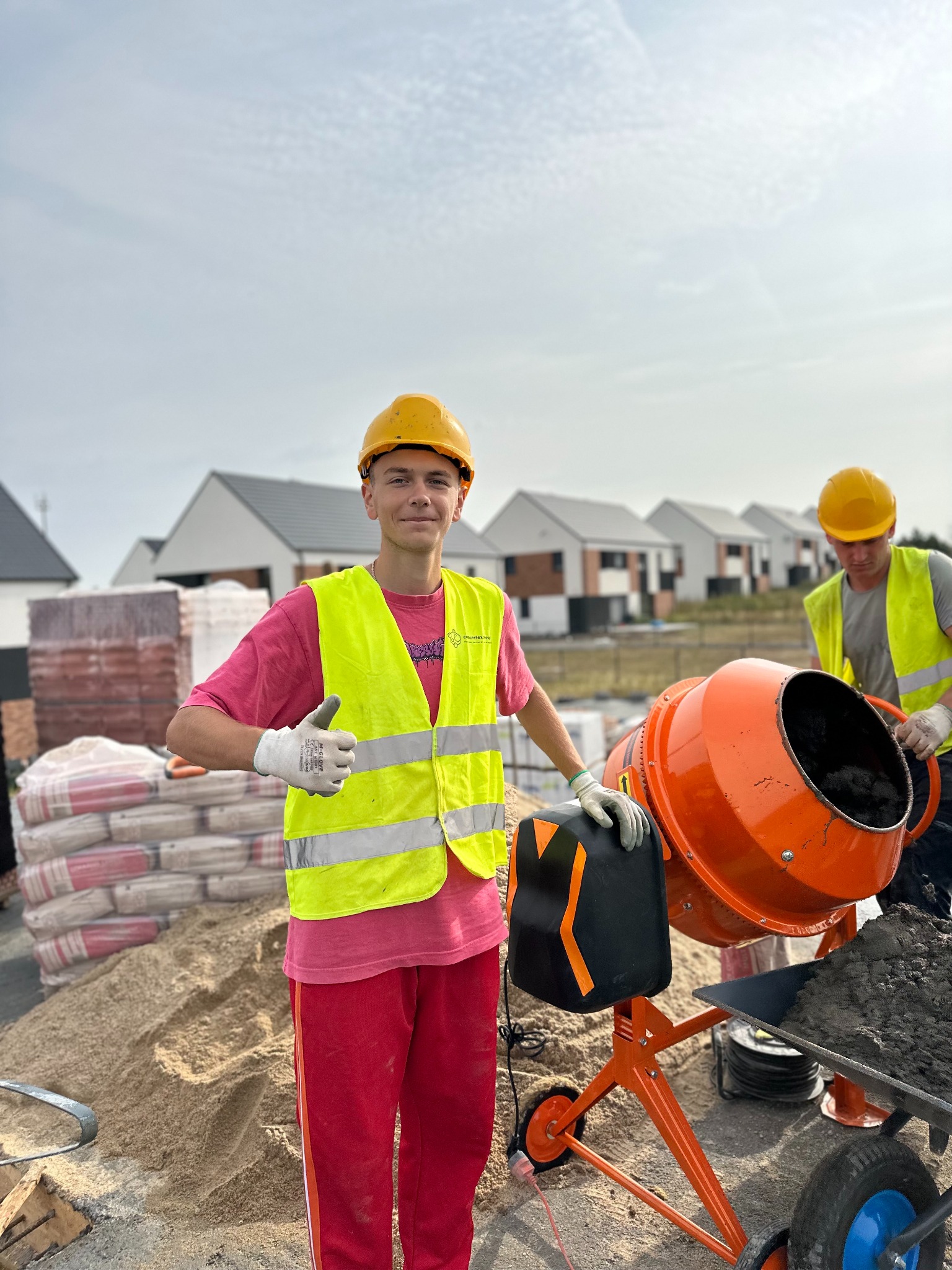Młody pracownik w żółtym kasku i kamizelce odblaskowej, z uśmiechem i uniesionym kciukiem, stoi obok pomarańczowej betoniarki na budowie domów jednorodzinnych.