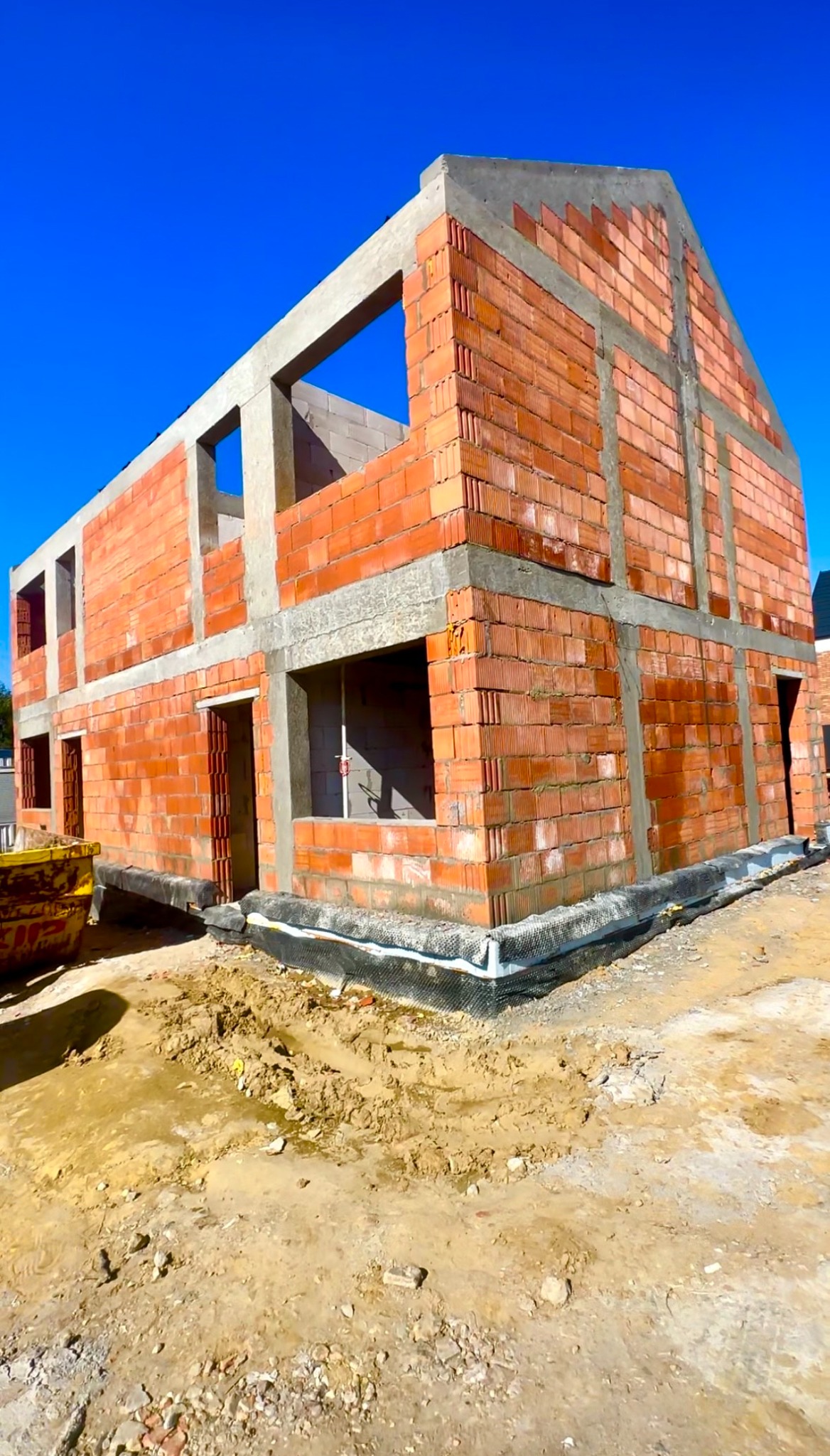 Surowy stan domu w trakcie budowy z czerwonej cegły, widoczne otwory okienne i drzwiowe, betonowe nadproża, teren budowy z błotem i kontenerem.