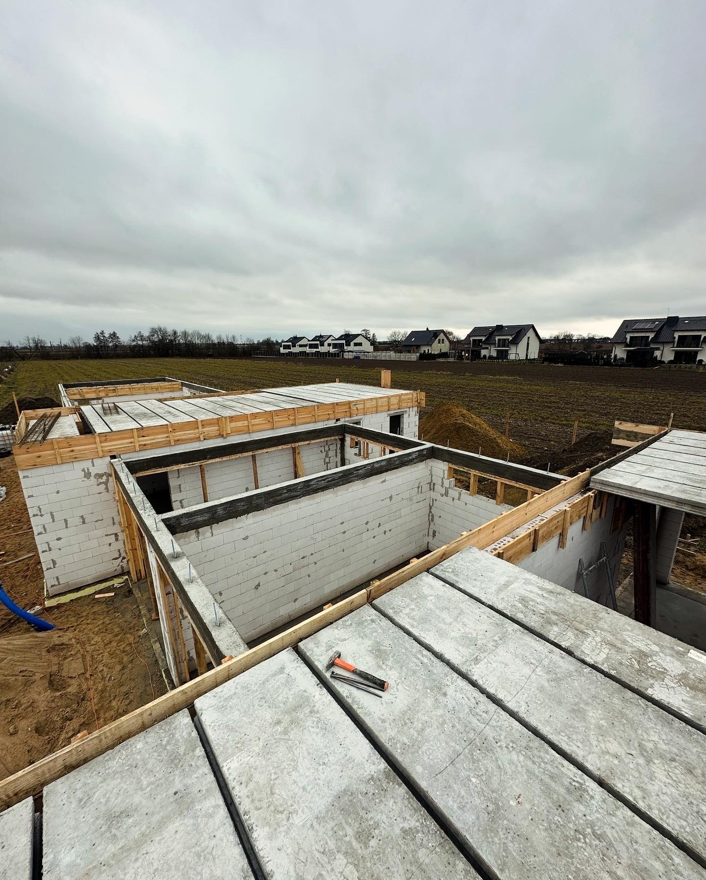 Budowa domu jednorodzinnego w trakcie wznoszenia ścian z bloczków betonowych i zalewania stropu, widoczne drewniane szalunki, zbrojenia i narzędzia na betonowych płytach stropowych, w tle zabudowa...