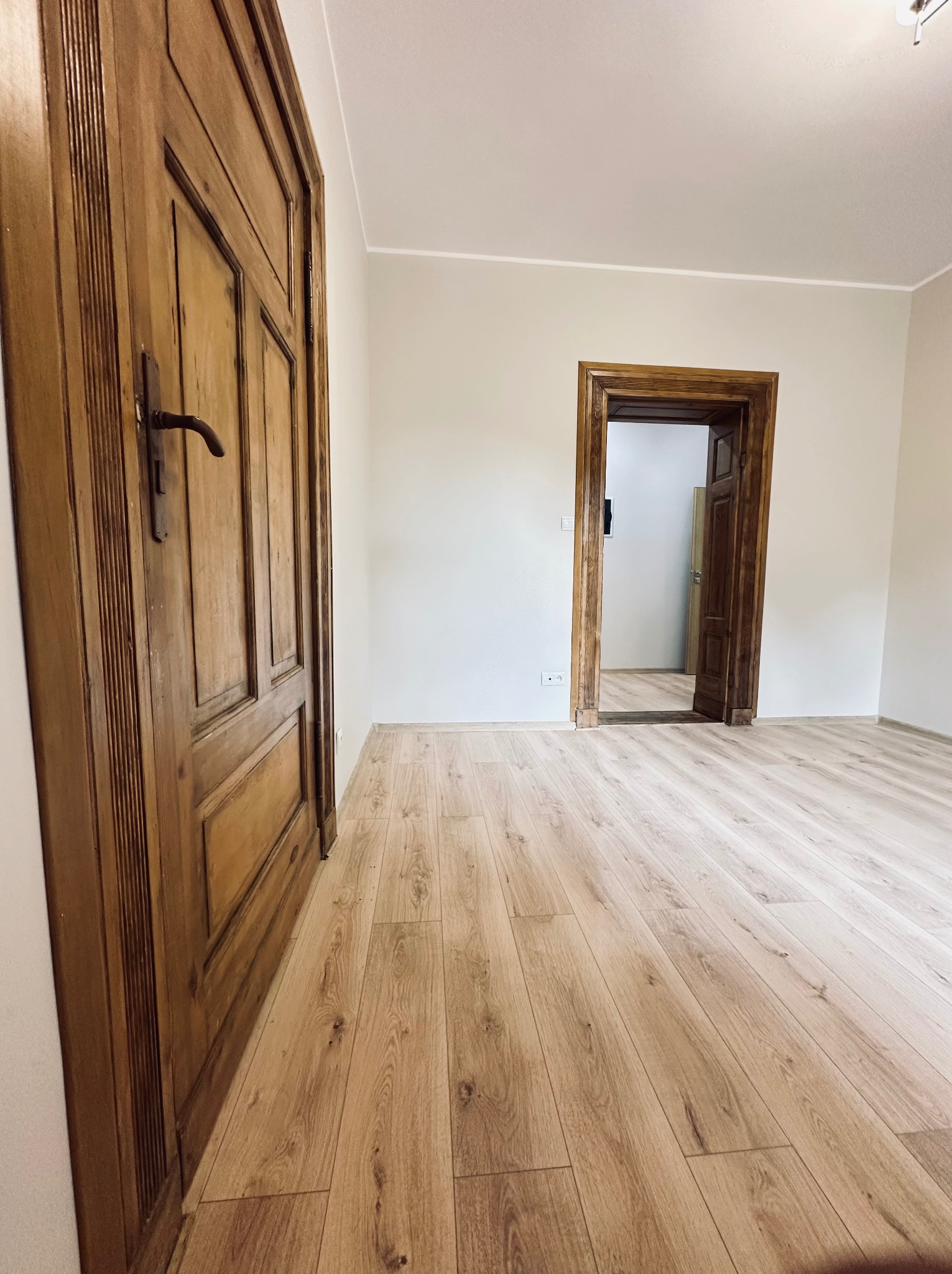 Jasne panele podłogowe w odnowionym pokoju z dwojgiem drewnianych drzwi, jedne zamknięte, drugie otwarte, ukazujące kolejne pomieszczenie. Świeżo pomalowane ściany.