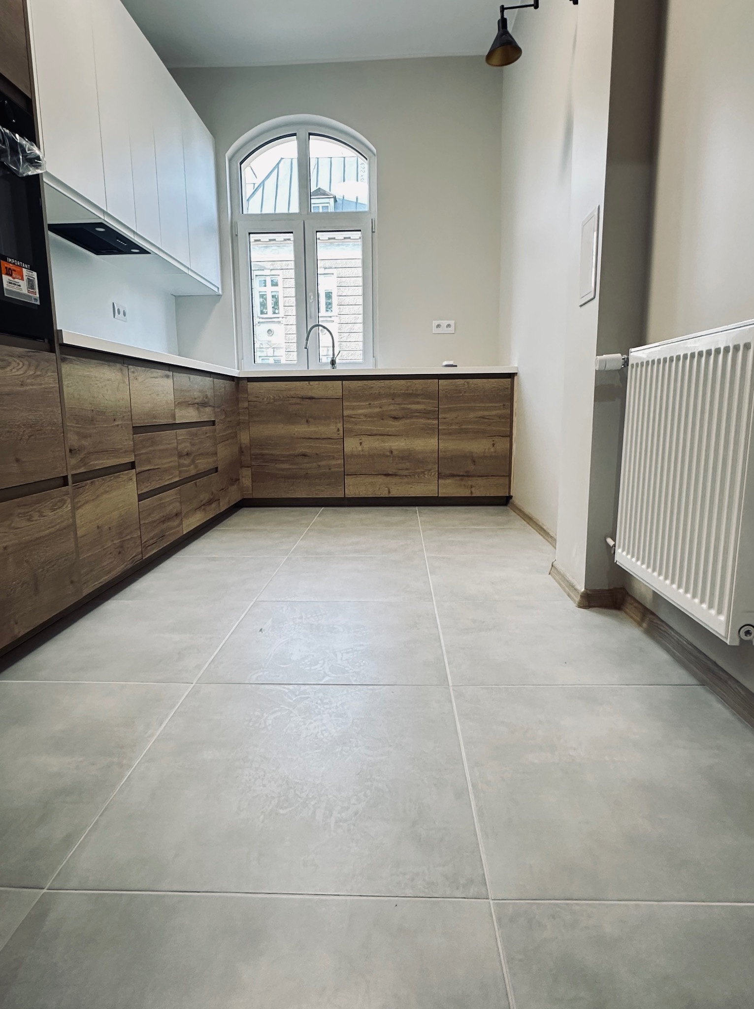 Jasna kuchnia po remoncie: szare płytki podłogowe, drewniane szafki z białymi górnymi szafkami, okno łukowe i grzejnik. Minimalistyczny design.