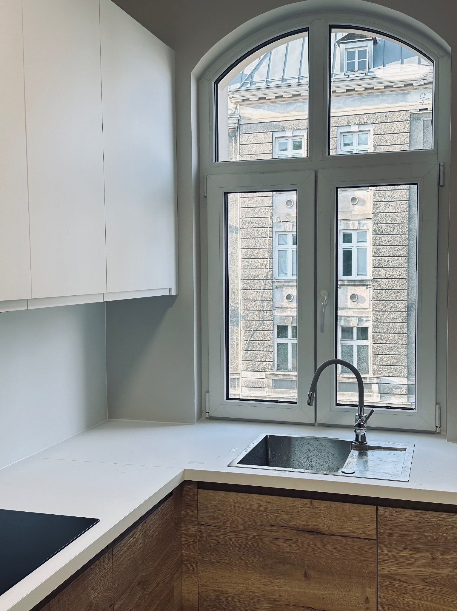 Nowoczesna kuchnia z drewnianymi szafkami i białym blatem, zlewem pod oknem w kamienicy. Minimalistyczny design, jasne wnętrze, widok na budynek z okna.