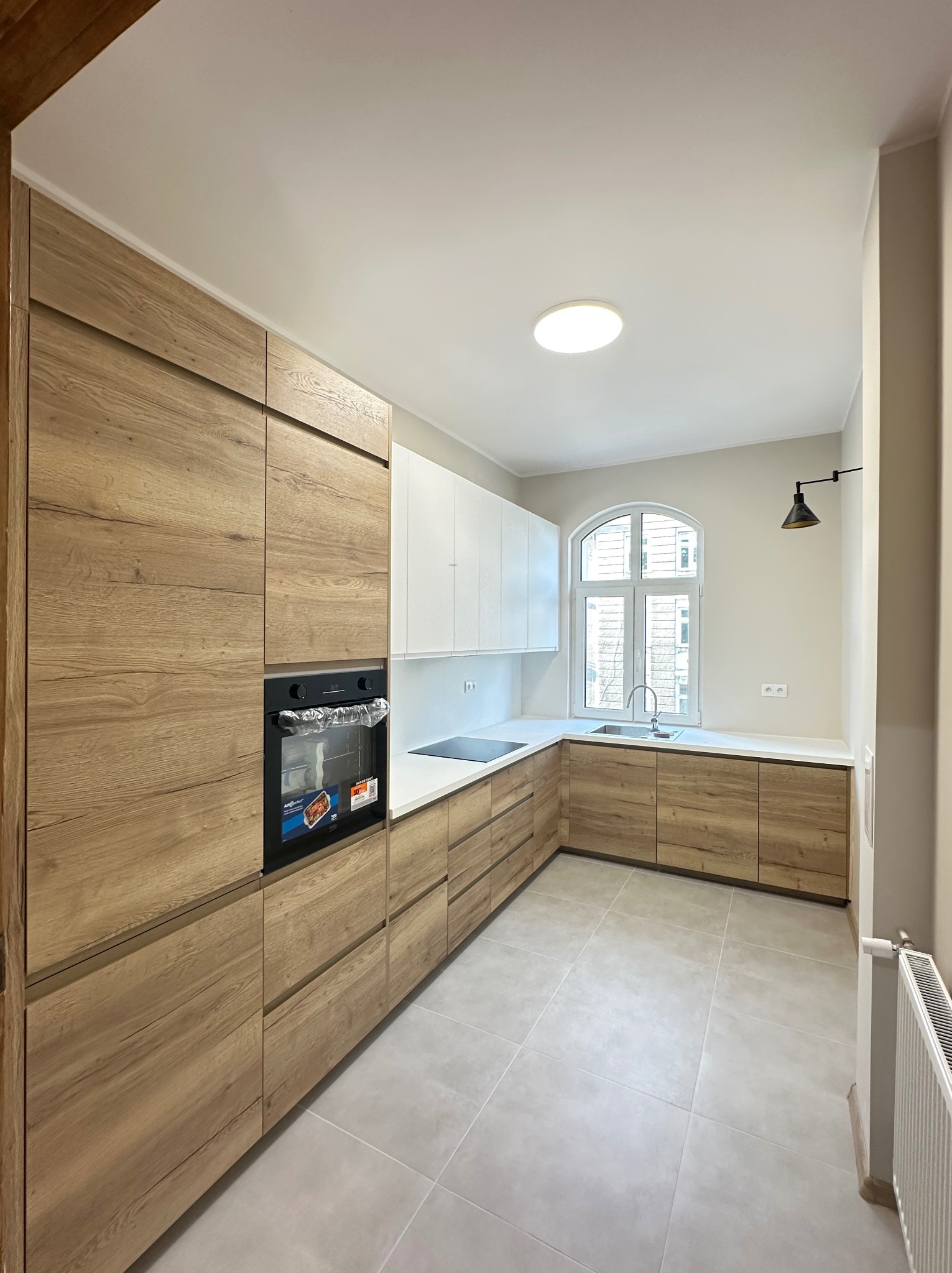 Jasna kuchnia po remoncie: drewniane szafki dolne, białe górne, okno z łukiem i szara podłoga. Widoczny piekarnik i czarna lampa ścienna.