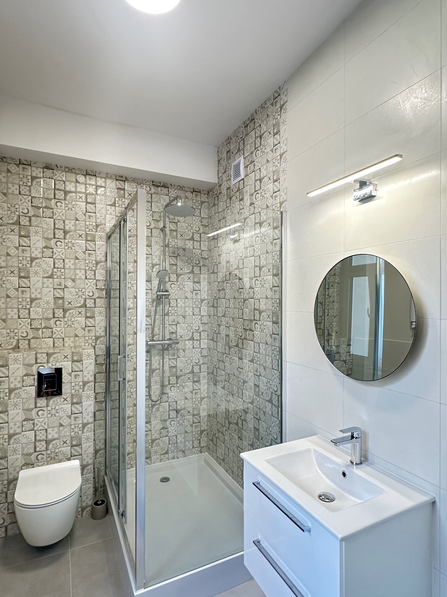 Nowoczesna łazienka po remoncie: kabina prysznicowa ze szklanymi drzwiami, umywalka z szafką, okrągłe lustro i toaleta w jasnych, neutralnych barwach.