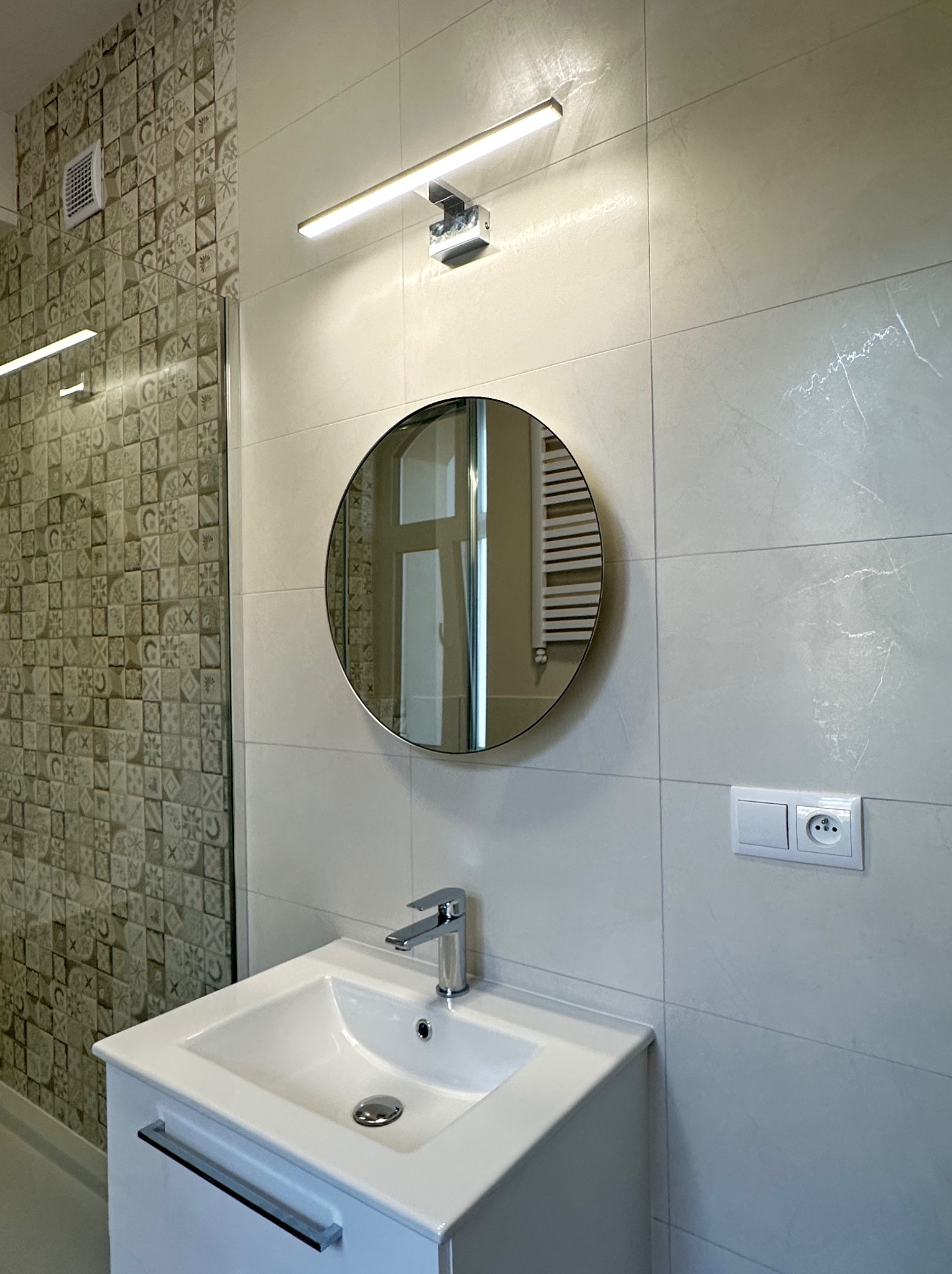 Nowoczesna łazienka po remoncie: biała umywalka z szafką, okrągłe lustro z podświetleniem LED i dekoracyjne płytki w strefie prysznica.