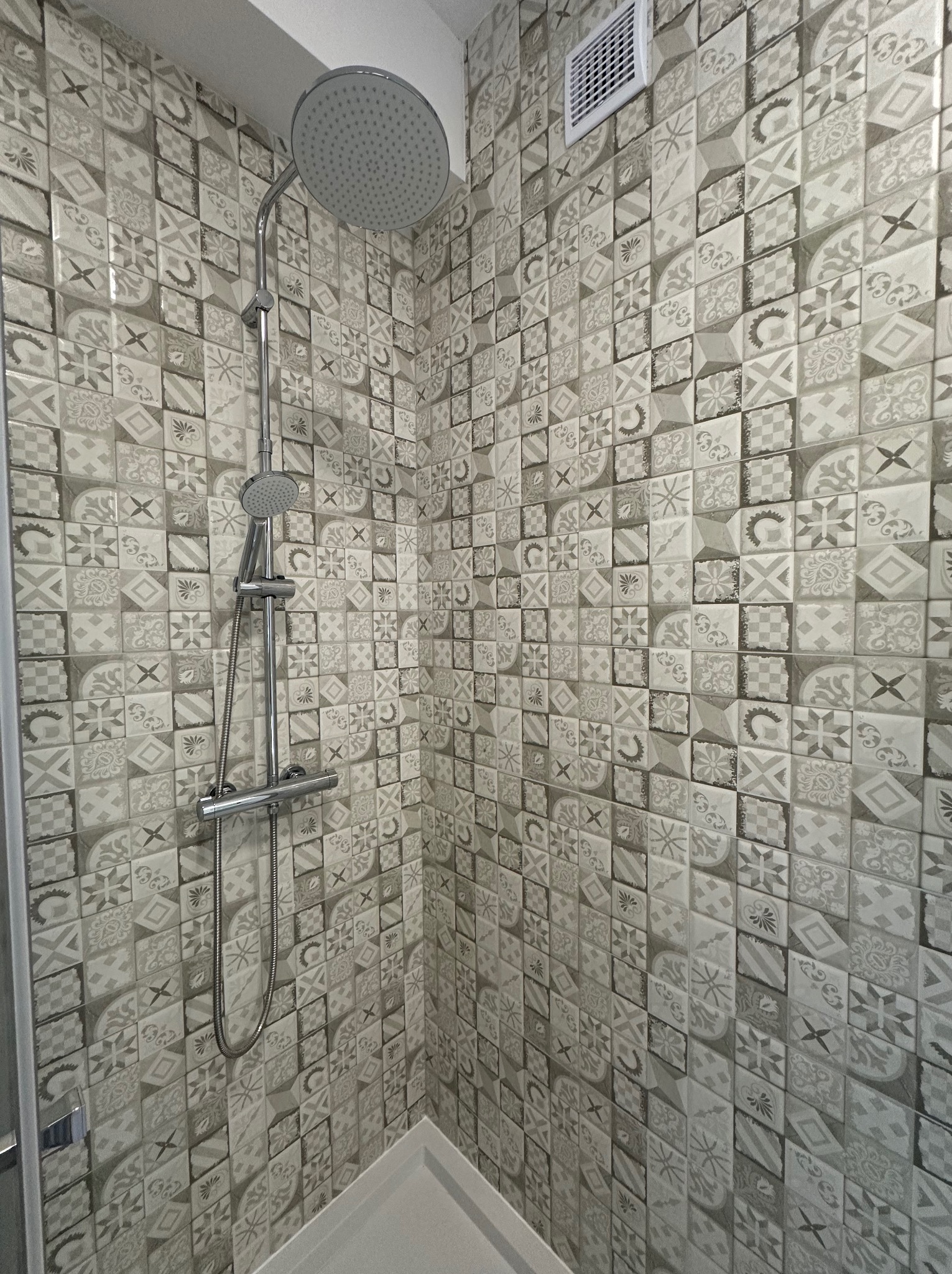 Wykończona kabina prysznicowa z mozaiką w odcieniach szarości. Widoczna bateria i deszczownica. Na ścianie kratka wentylacyjna. Nowoczesny design.