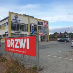 Fasada budynku firmy Amicus z reklamą zewnętrzną 'Drzwi' na czerwonym banerze i szyldami 'Rewolucje Łazienkowe' oraz 'Drzwi Podłogi', widok na parking i samochody