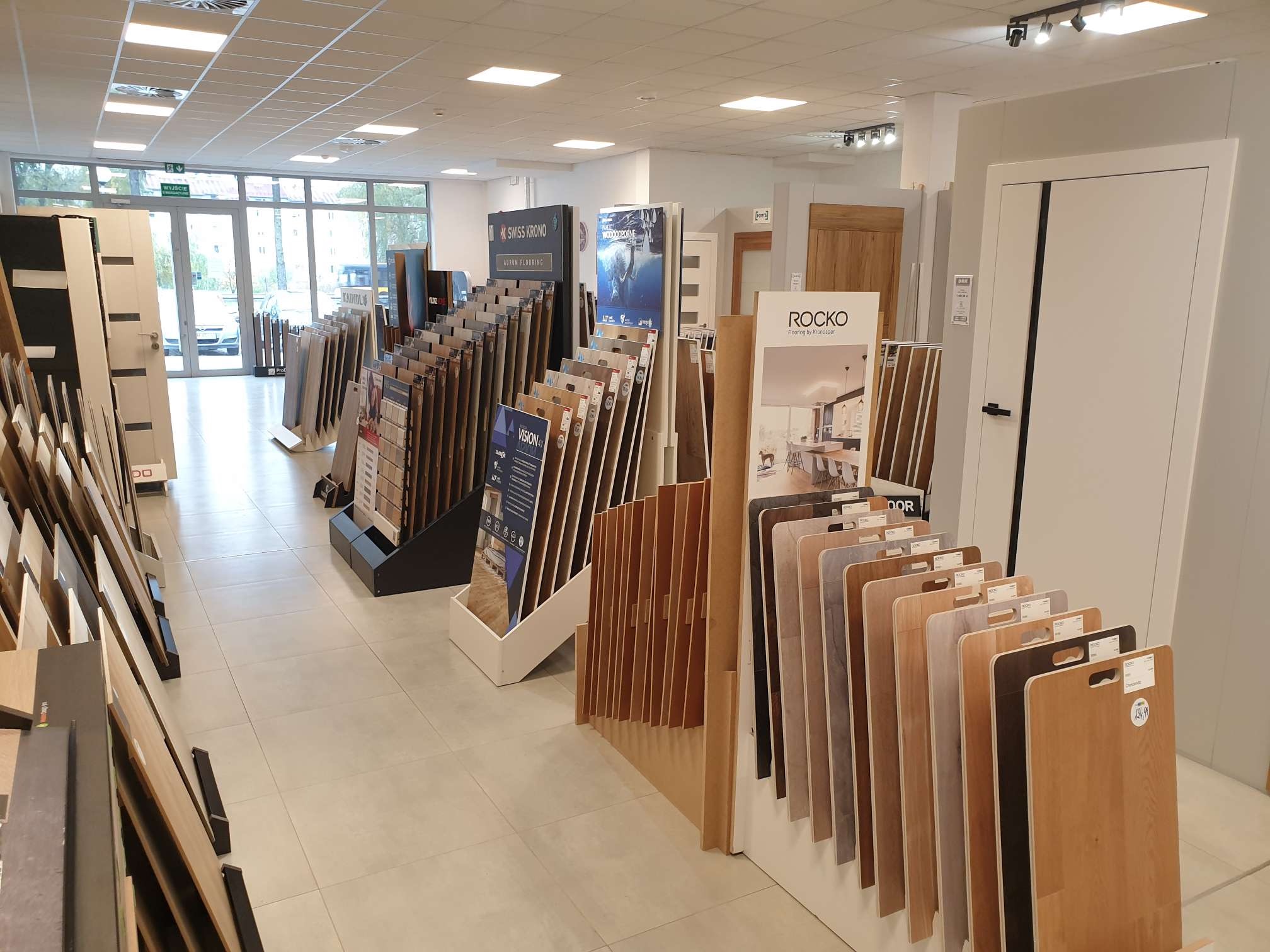 Ekspozycja różnorodnych paneli podłogowych i drzwi w salonie sprzedaży, widoczne próbki paneli w odcieniach drewna i szarości oraz drzwi w różnych stylach, oświetlenie sufitowe.