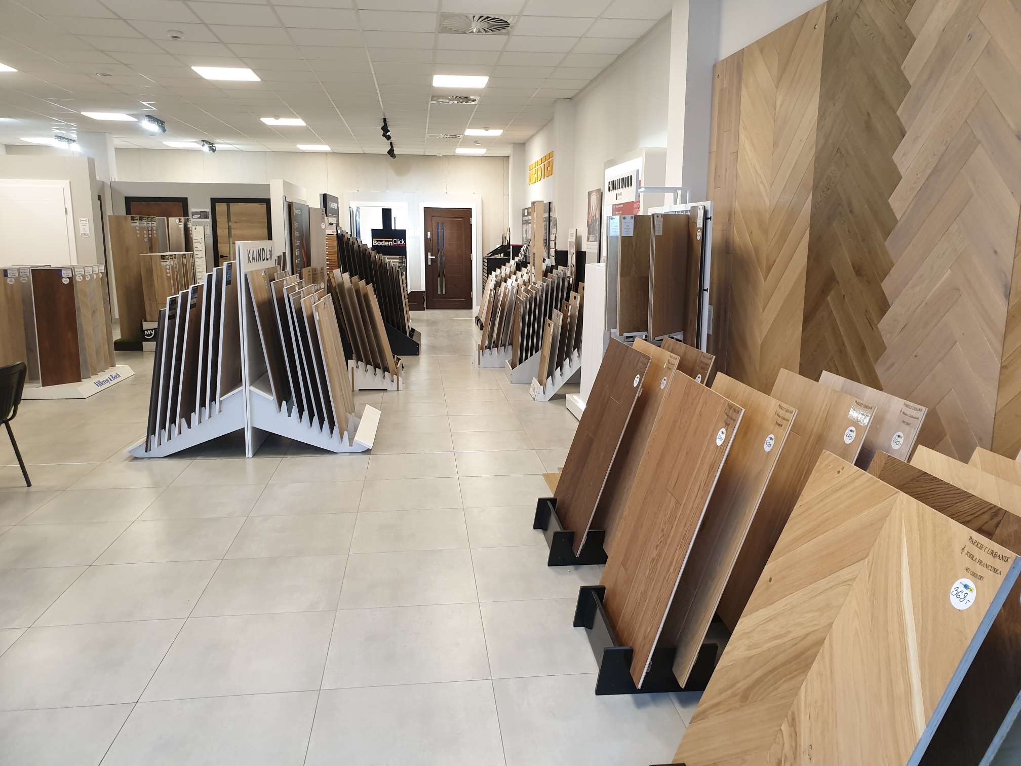 Ekspozycja różnorodnych paneli i parkietów w salonie sprzedaży, widoczne wzory jodełki francuskiej i tradycyjne deski, oświetlenie sufitowe, płytki podłogowe w tle.