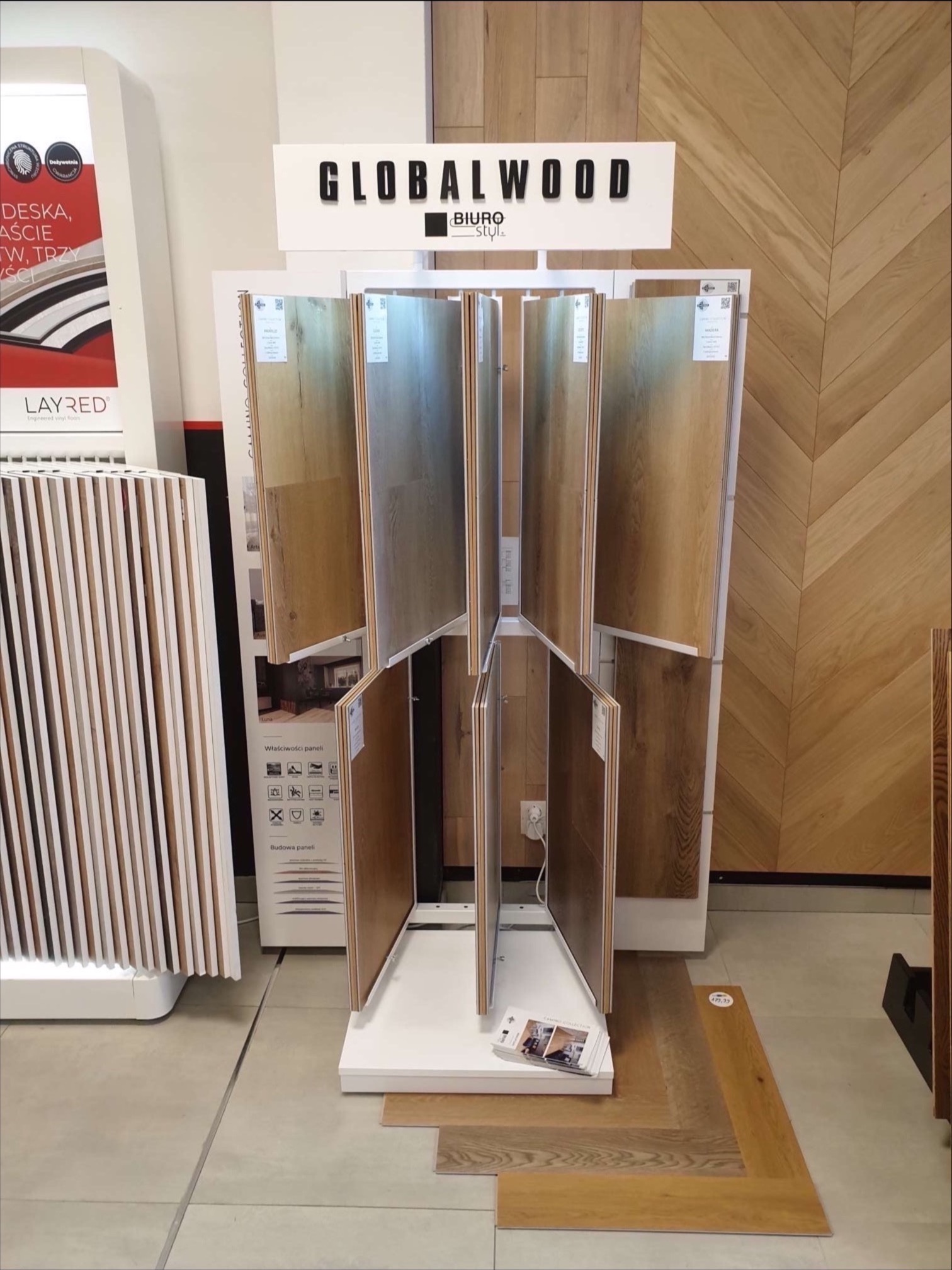 Ekspozycja próbek paneli podłogowych w salonie, różne odcienie drewna na obrotowym stojaku, w tle dekoracyjna ściana z parkietu ułożonego w jodełkę, widoczne materiały informacyjne i logo producenta.