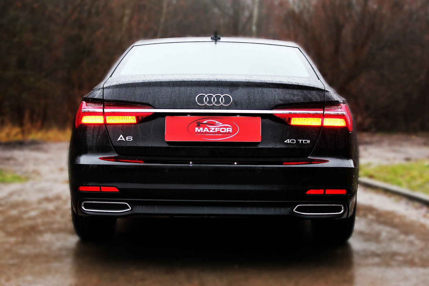 Tył czarnego Audi A6 40 TDI z włączonymi światłami, na mokrej nawierzchni, z czerwoną tabliczką z logo firmy na klapie bagażnika.