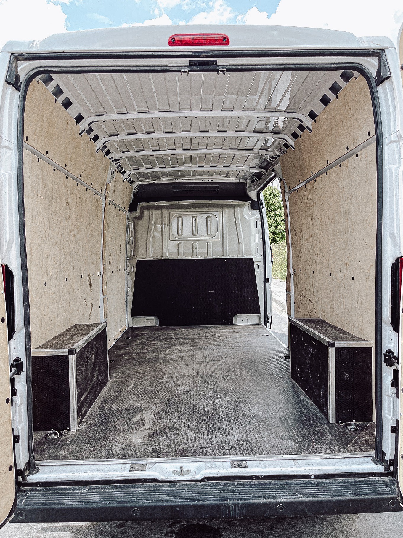 Wnętrze białego furgonetki wyłożone sklejką, z dwoma czarnymi skrzynkami po bokach, widok z tyłu przez otwarte drzwi.