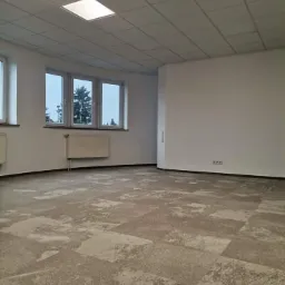 Biuro z nową, wzorzystą wykładziną dywanową w odcieniach szarości i beżu, widok na grzejniki pod oknami i sufit z kasetonami oraz klimatyzacją.