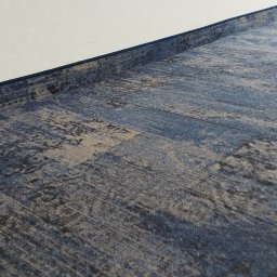 Gold Floor - Niebiesko-szara wykładzina dywanowa z wzorem abstrakcyjnym, ułożona przy białej ścianie z widoczną listwą przypodłogową.