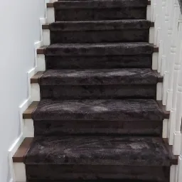 Schody w domu pokryte brązową wykładziną dywanową, białe listwy przypodłogowe i drewniane stopnice.