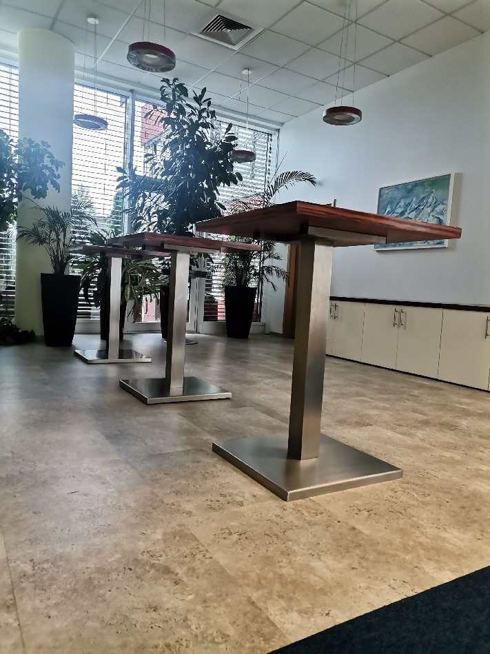 Wnętrze biurowe z jasną, wzorzystą posadzką imitującą kamień, na której stoją trzy wysokie stoliki z ciemnymi drewnianymi blatami i metalowymi nogami, w tle rośliny w donicach i okna.