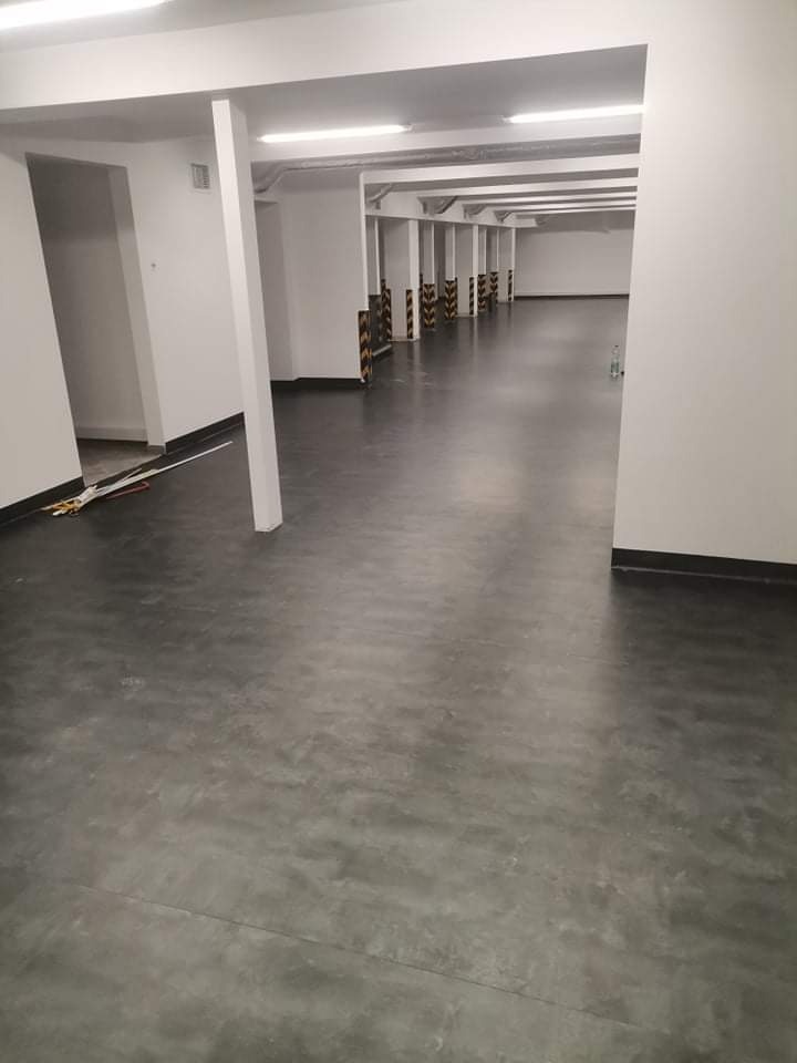 Szara, gładka podłoga w hali magazynowej lub garażu podziemnym z białymi ścianami i słupami. Na słupach widoczne czarno-żółte odbojniki. W tle widoczna butelka wody.