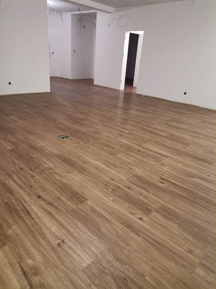 Nowo położona, jasnobrązowa podłoga w pustym, jasnym pomieszczeniu z białymi ścianami i widocznymi otworami wentylacyjnymi w podłodze.