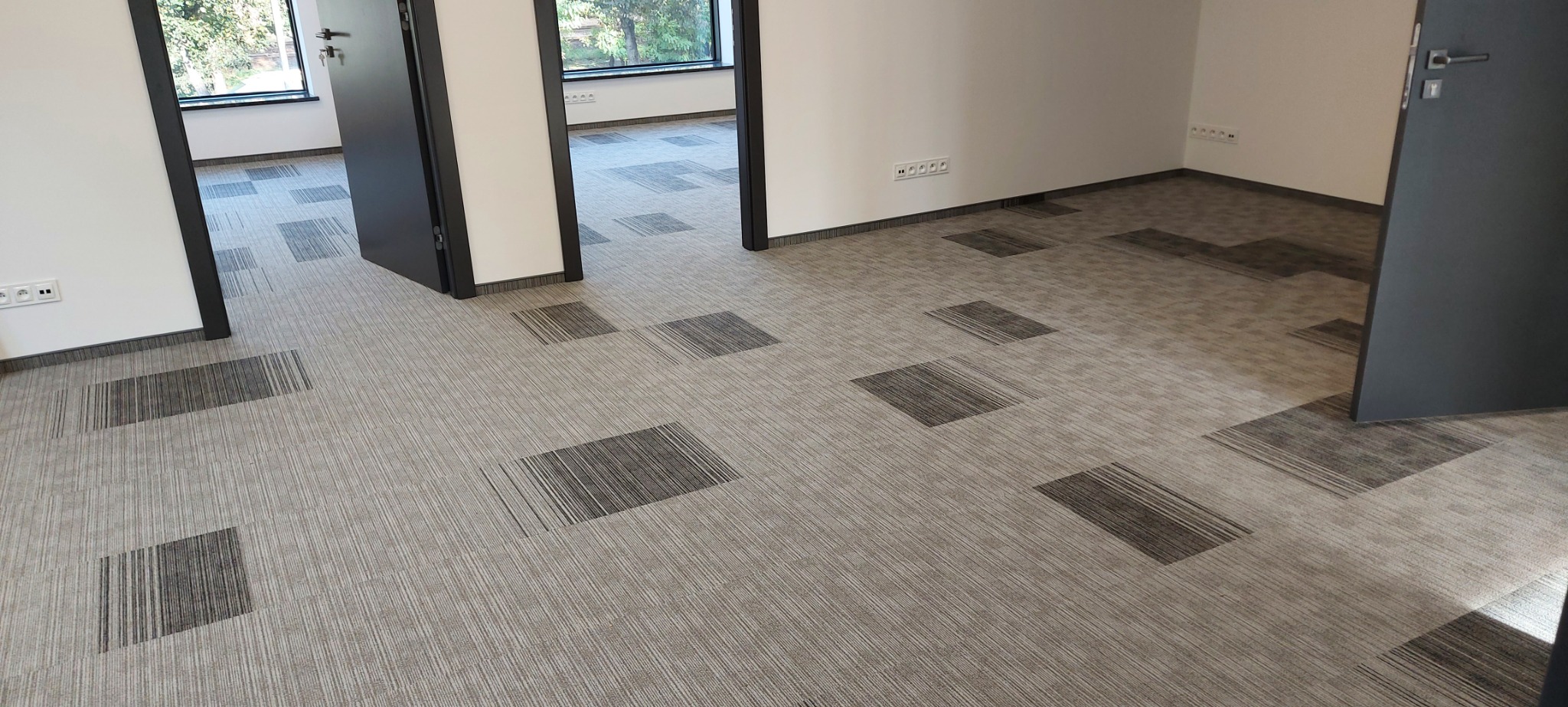 Beżowa wykładzina dywanowa w kwadraty z szarymi paskami ułożona w biurze z białymi ścianami i czarnymi futrynami drzwi.