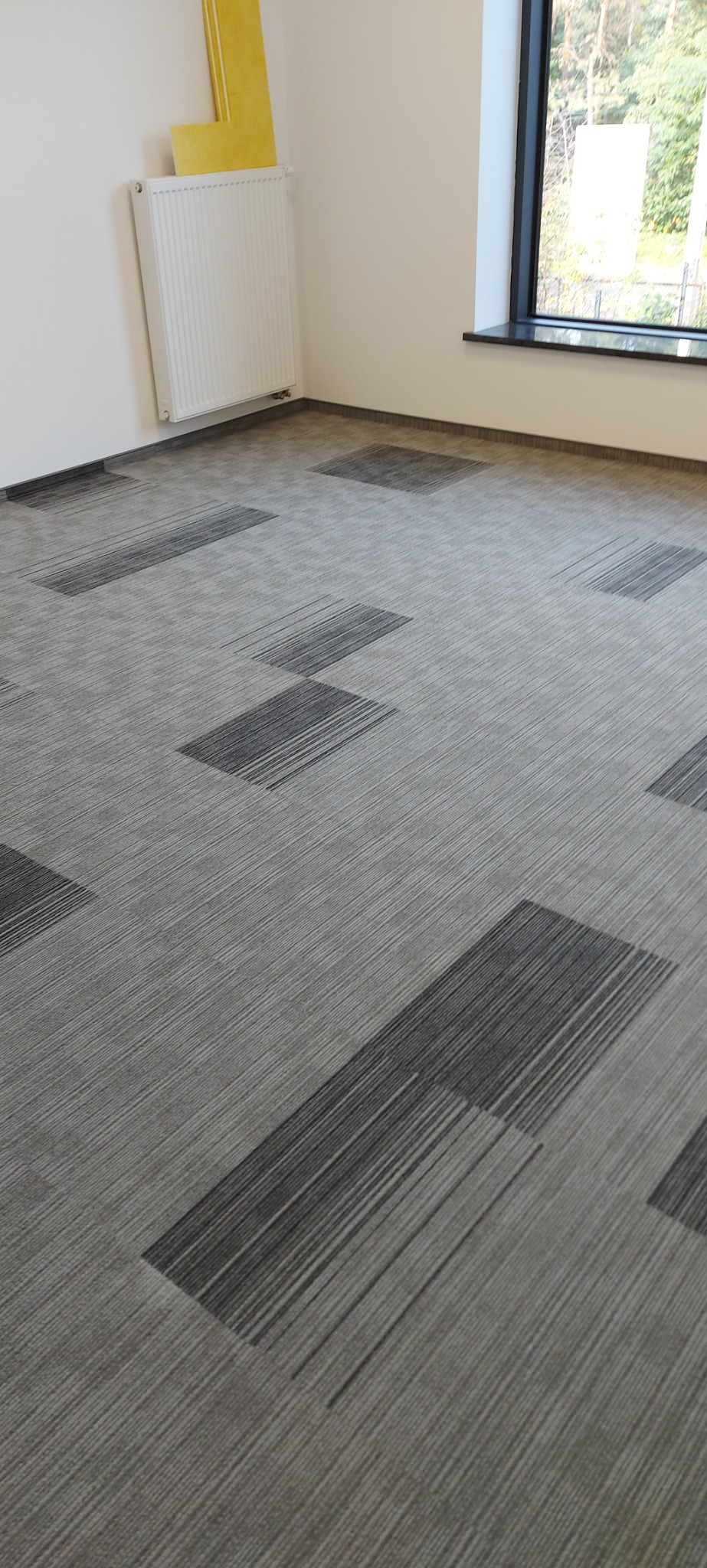 Szara wykładzina dywanowa w płytkach z ciemniejszymi, prostokątnymi wzorami w nowym biurze, widok na grzejnik i okno z widokiem na drzewa.