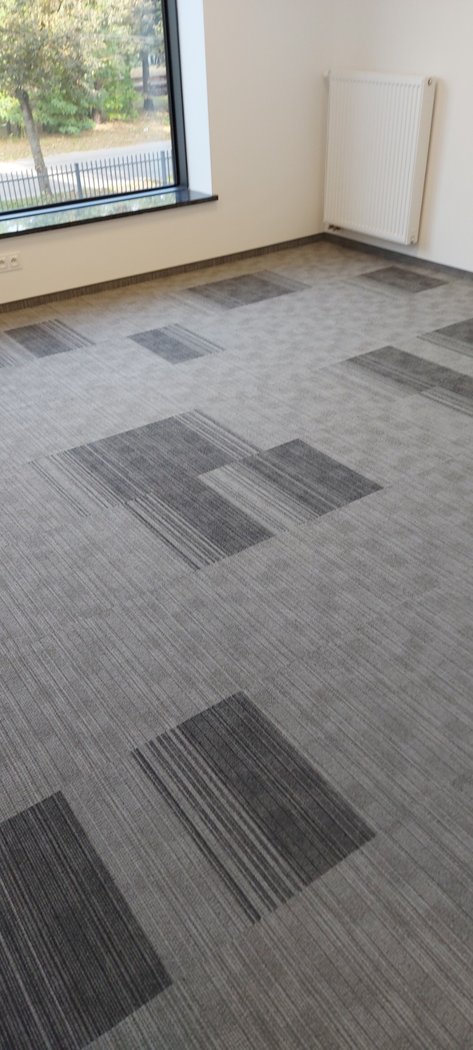 Szara wykładzina dywanowa w kwadraty i paski, ułożona w pustym, jasnym pomieszczeniu z oknem i grzejnikiem.