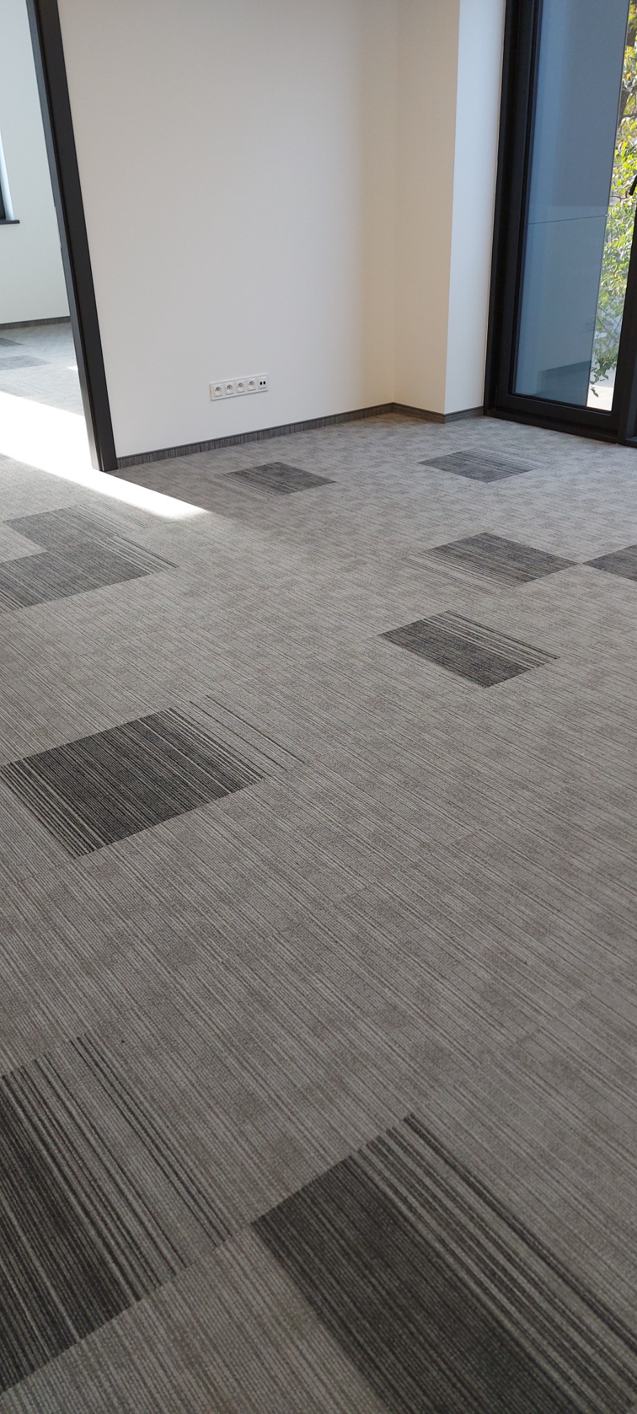 Szara wykładzina dywanowa w kwadraty z widocznymi liniami, ułożona w pustym pomieszczeniu biurowym z białymi ścianami i oknami.