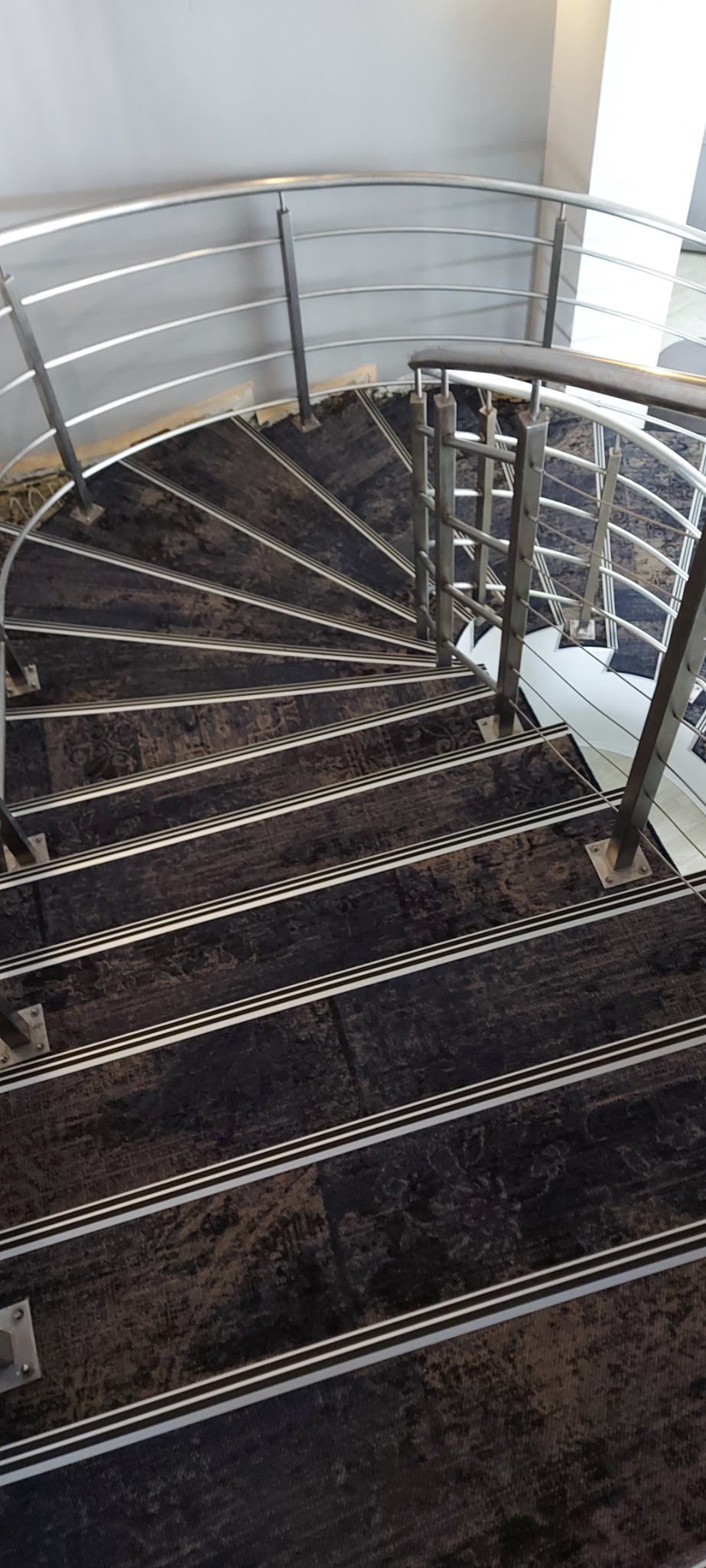 Spiralne schody pokryte ciemną wykładziną z jasnymi paskami antypoślizgowymi i metalową balustradą, widok z góry.
