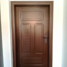 Odnowione, ciemnobrązowe drzwi wewnętrzne z trzema panelami, z delikatnym połyskiem, osadzone w białej ścianie z widoczną klamką w stylu retro.