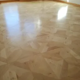 Wypolerowana parkietowa podłoga ułożona we wzór gwiazdy, widoczne refleksy światła słonecznego wpadającego przez okno, białe ściany i grzejnik w tle.