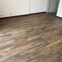 Ułożone, brązowe płytki podłogowe imitujące drewno w nowym, jasnym pomieszczeniu z białymi ścianami i szarą listwą przypodłogową.