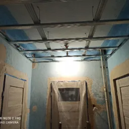 Metalowa konstrukcja sufitu podwieszanego z płyt kartonowo-gipsowych, widok w trakcie montażu, ściany w stanie surowym, zabezpieczone drzwi folią ochronną, widoczne kable i oświetlenie tymczasowe.