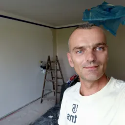 Mężczyzna z niebieskimi oczami robi selfie na tle pokoju w trakcie odświeżania, widoczna drabina, puszka z farbą i niebieska folia ochronna na suficie.