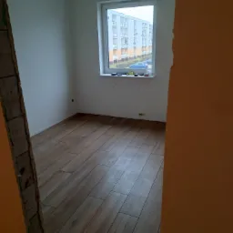 Pokój po remoncie z nową podłogą z paneli, widok przez futrynę, za oknem blok mieszkalny.