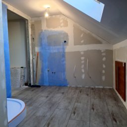 Usługi Remontowo-Budowlane Arkadiusz Witek - Poddasze w trakcie prac wykończeniowych: ściany z płyt kartonowo-gipsowych, częściowo pokryte gruntem, widoczne instalacje, ułożona podłoga z paneli imitujących drewno, kabina prysznicowa w rogu.