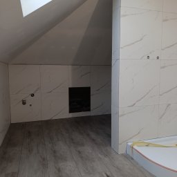 Usługi Remontowo-Budowlane Arkadiusz Witek - Wykończona łazienka na poddaszu ze skośnym sufitem, wyłożona białymi płytkami imitującymi marmur, z drewnopodobną podłogą i przygotowanym miejscem na prysznic.