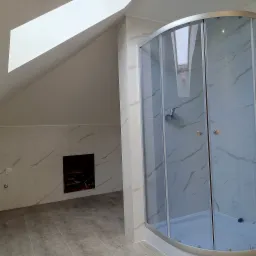 Wykończona łazienka na poddaszu ze skośnym sufitem i oknem dachowym, kabina prysznicowa z marmurowymi płytkami, szara podłoga z paneli, widoczny otwór rewizyjny.