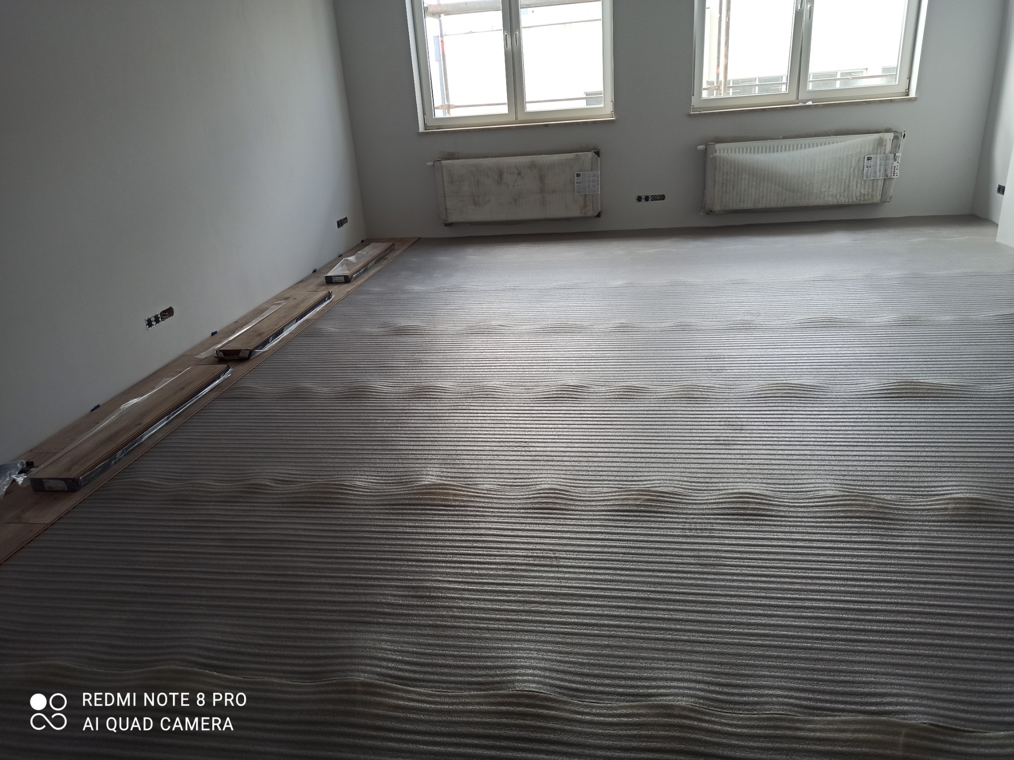 Przygotowanie do układania paneli: podkład falisty na podłodze, paczki paneli przy ścianie, grzejniki zabezpieczone folią, surowe ściany z widocznymi gniazdkami.