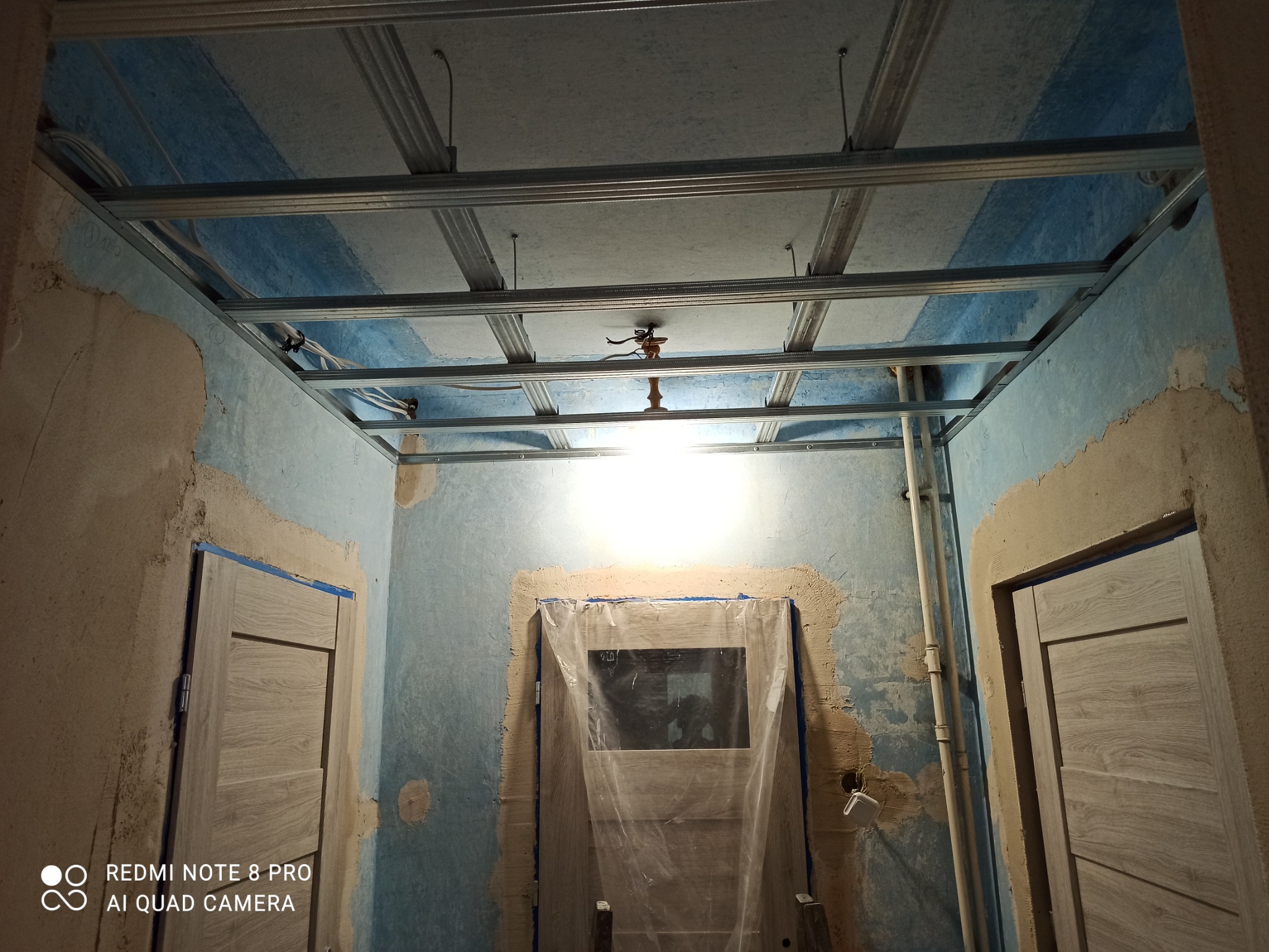 Metalowa konstrukcja sufitu podwieszanego z płyt kartonowo-gipsowych, widok w trakcie montażu, ściany w stanie surowym, zabezpieczone drzwi folią ochronną, widoczne kable i oświetlenie tymczasowe.