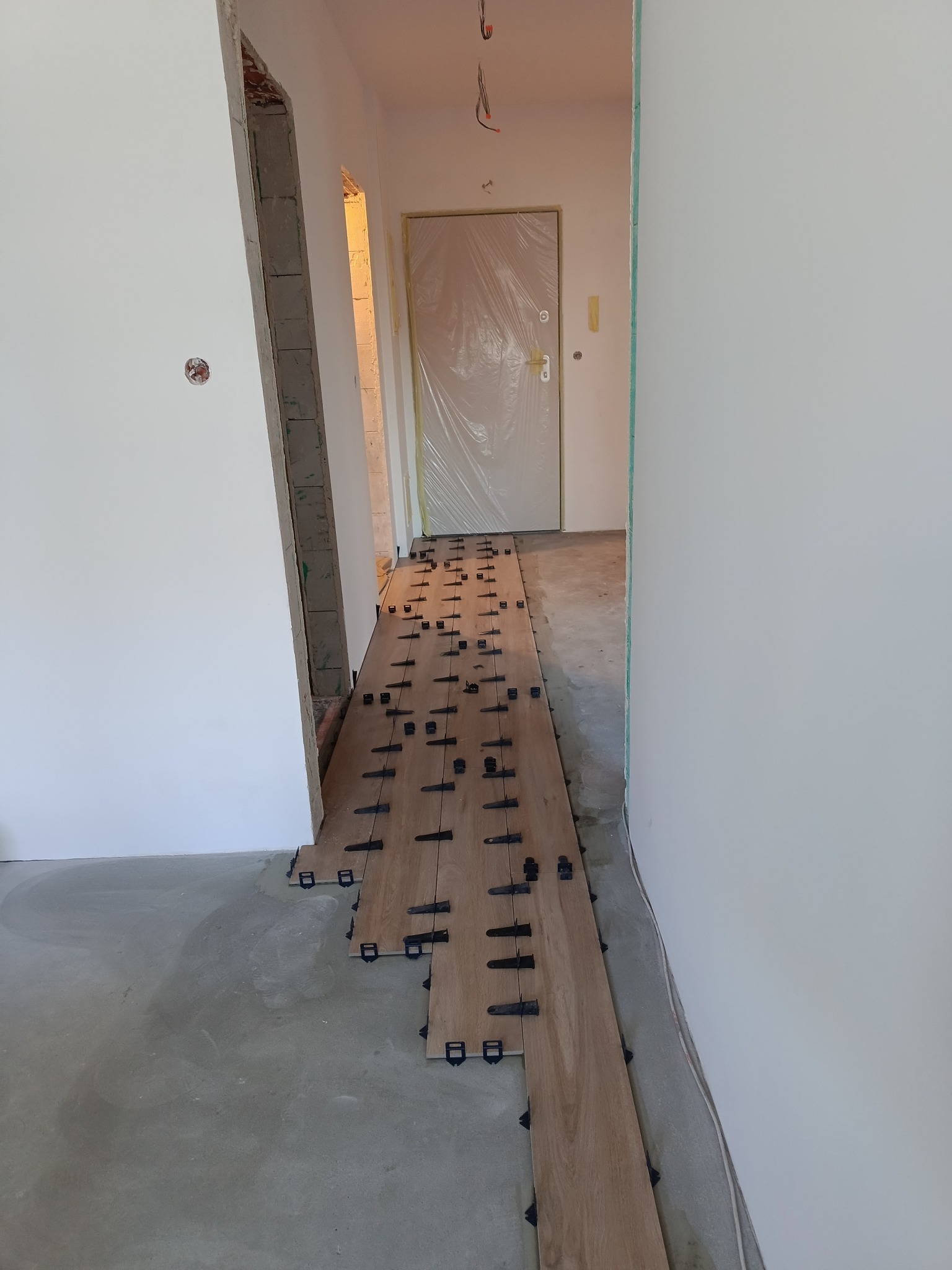 Fragment korytarza w trakcie układania paneli podłogowych z użyciem klipsów poziomujących, widoczne surowe ściany w trakcie remontu.