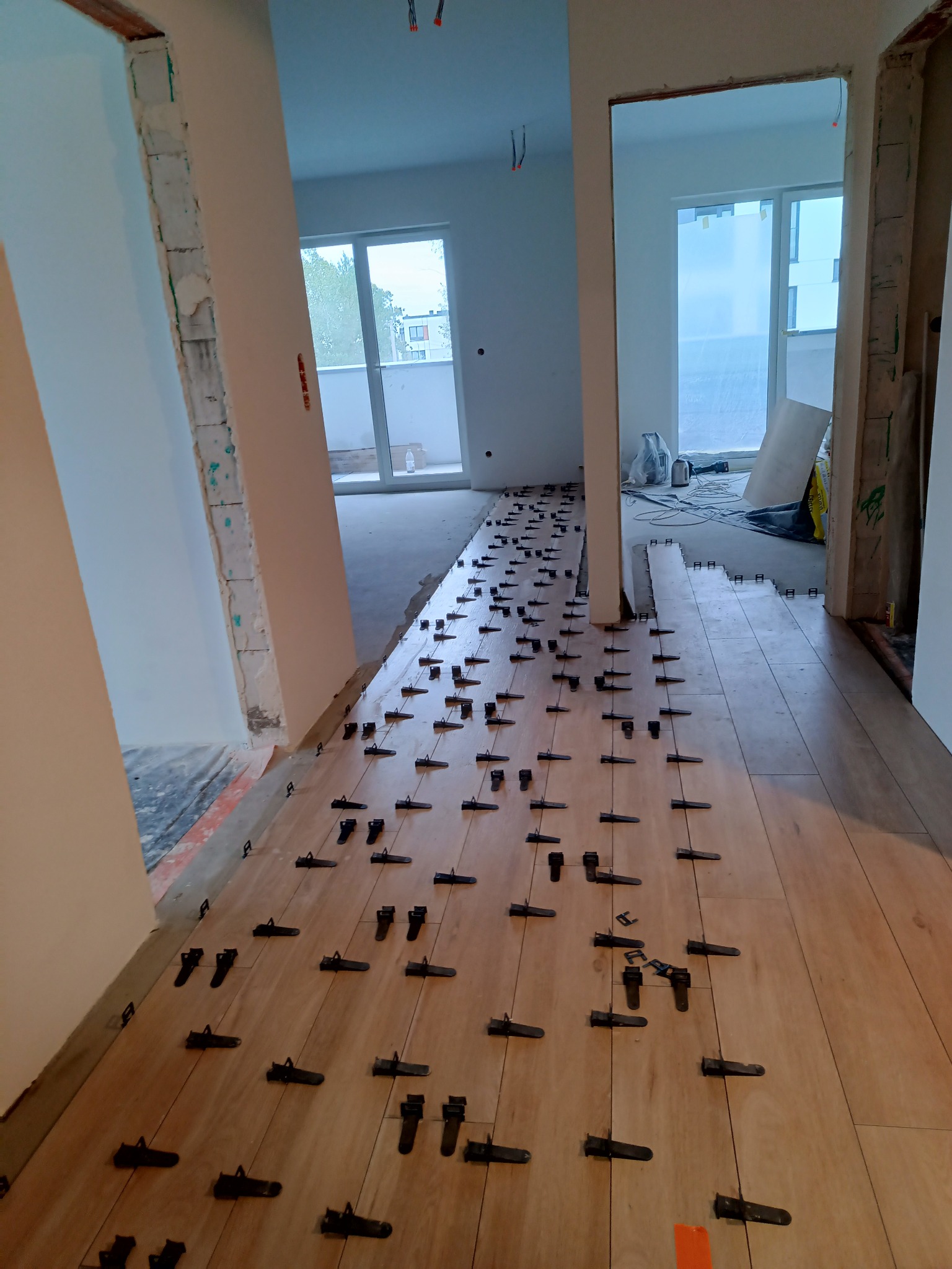 Montaż paneli podłogowych w długim korytarzu, z widocznymi czarnymi klipsami poziomującymi, surowe ściany w tle i okna z widokiem na zewnątrz.