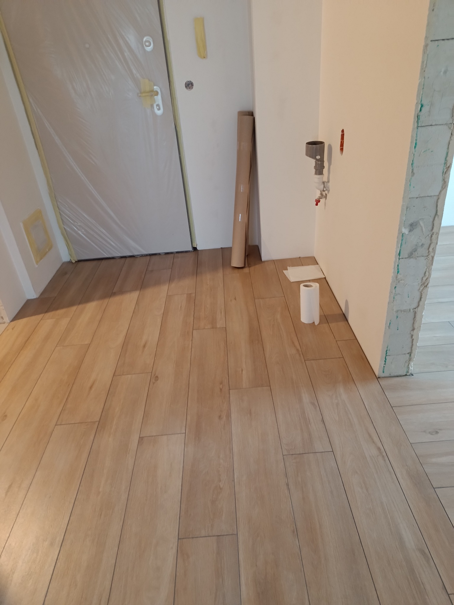 Ułożone panele podłogowe w odcieniach drewna, widoczna zabezpieczona folią drzwi wejściowe i fragment ściany z instalacją hydrauliczną.