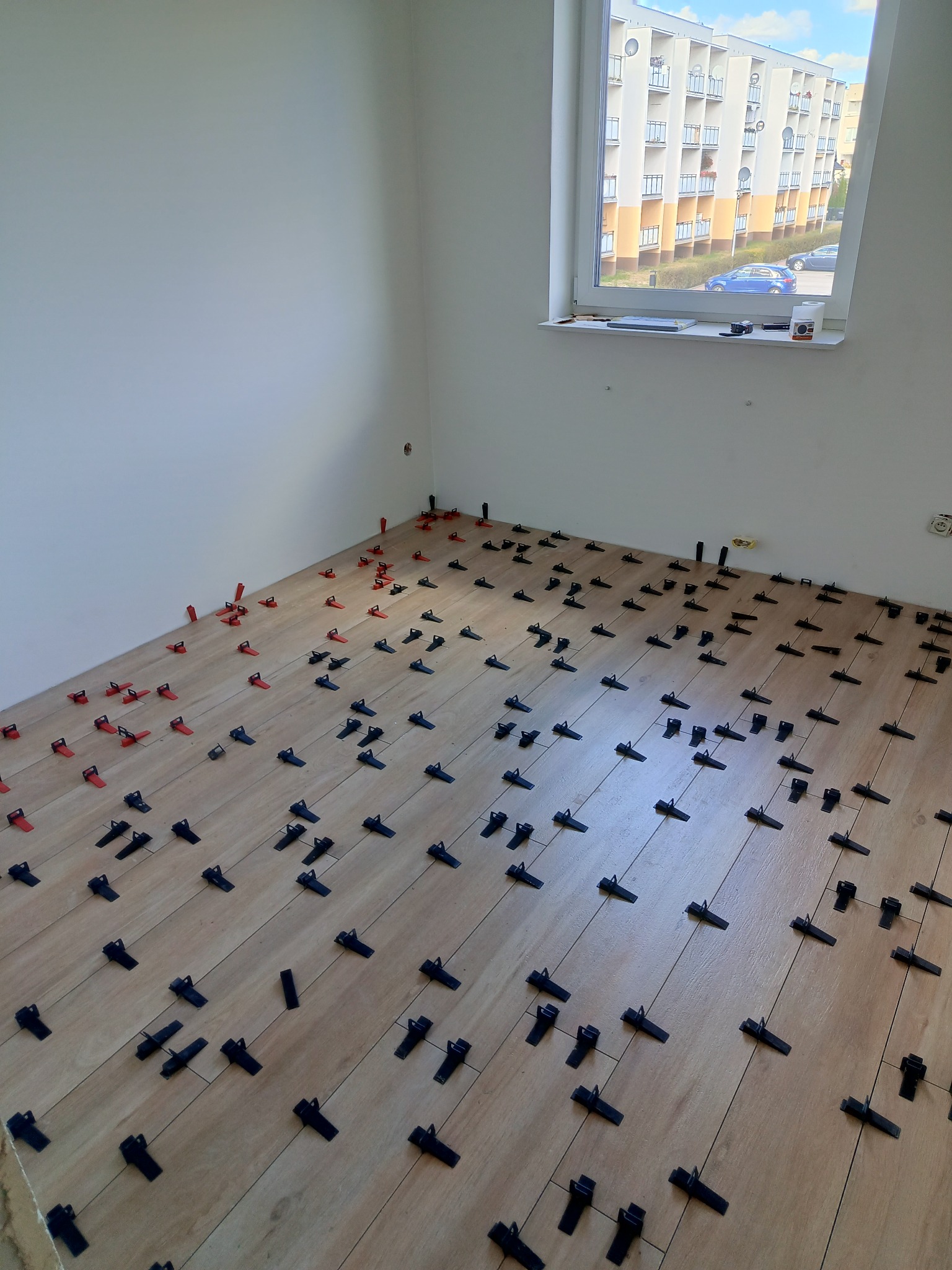 Podłoga z paneli ułożona z użyciem klipsów poziomujących, widok na okno z budynkiem mieszkalnym w tle.