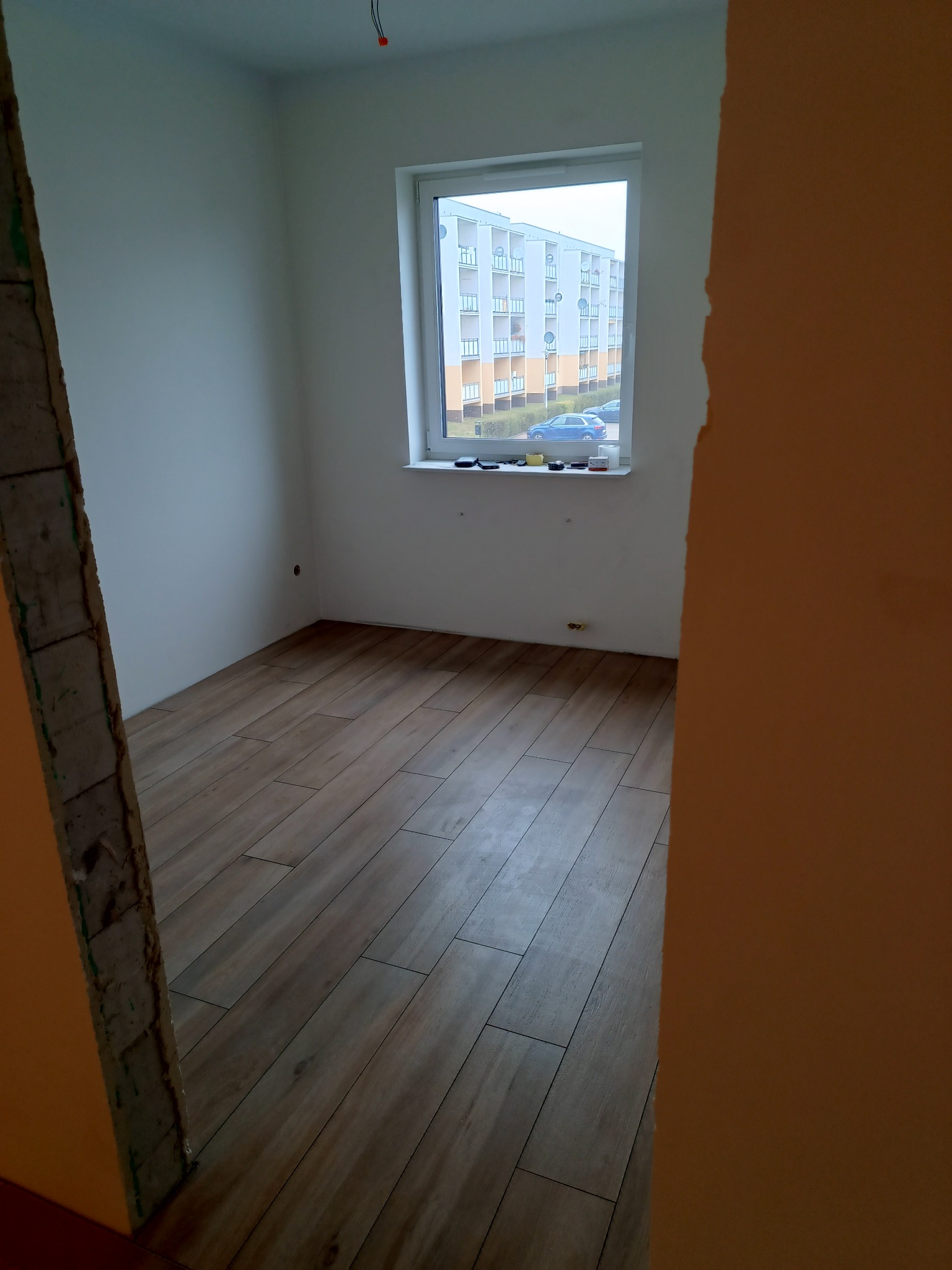 Pokój po remoncie z nową podłogą z paneli, widok przez futrynę, za oknem blok mieszkalny.