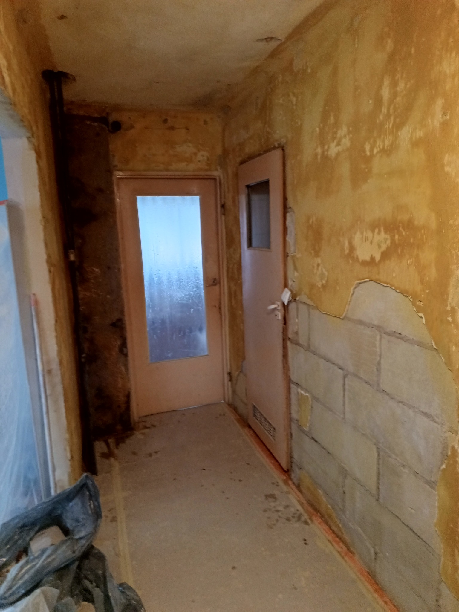 Korytarz w trakcie prac przygotowawczych do remontu, widoczne odsłonięte cegły pod warstwą starego tynku, drzwi z szybą, ślady wilgoci i rura przy suficie.