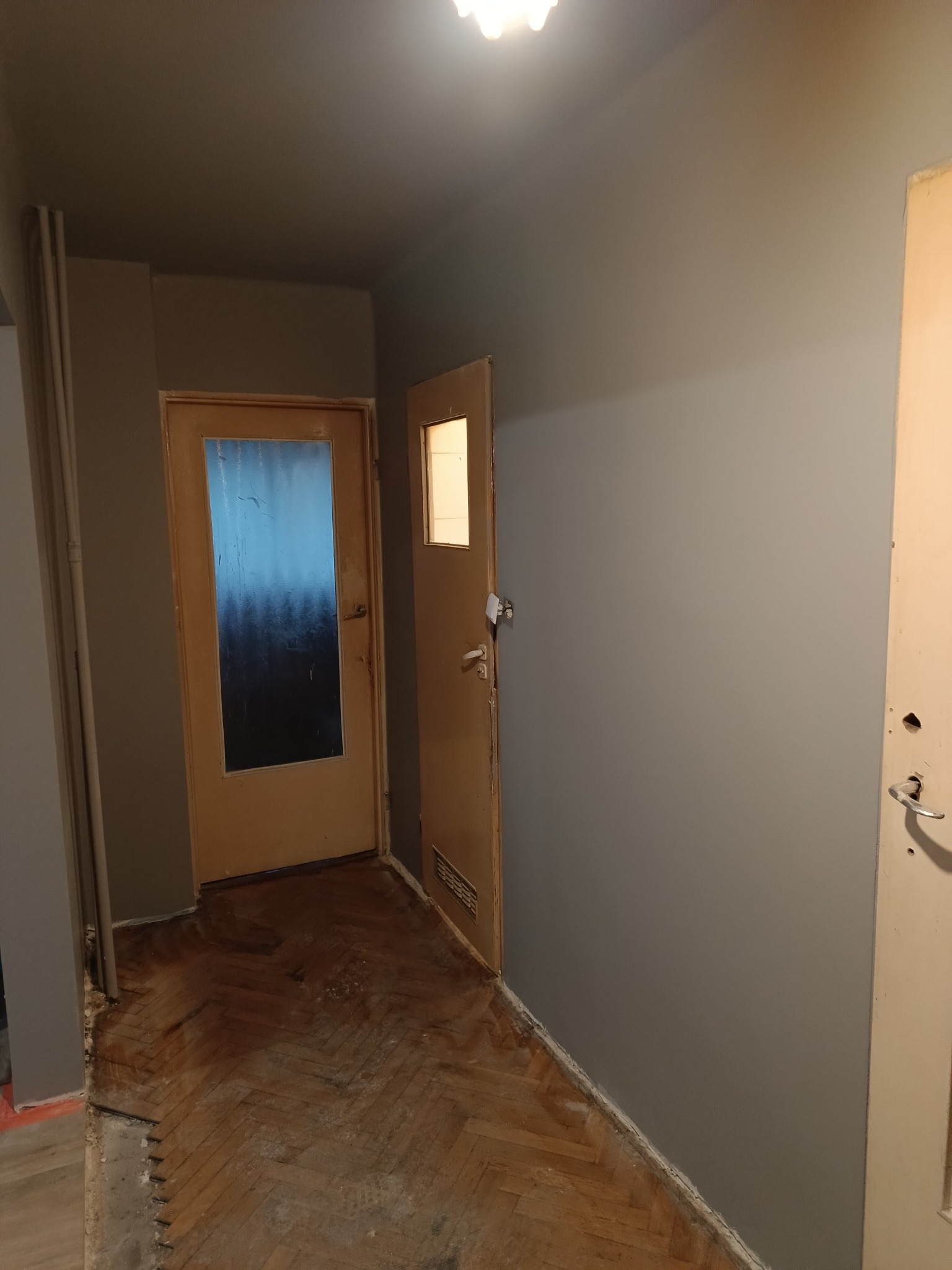 Korytarz w trakcie odświeżania, ściana pomalowana na szaro, widoczna stara drewniana podłoga w jodełkę częściowo odsłonięta, drzwi drewniane z szybą i drugie drzwi otwarte.