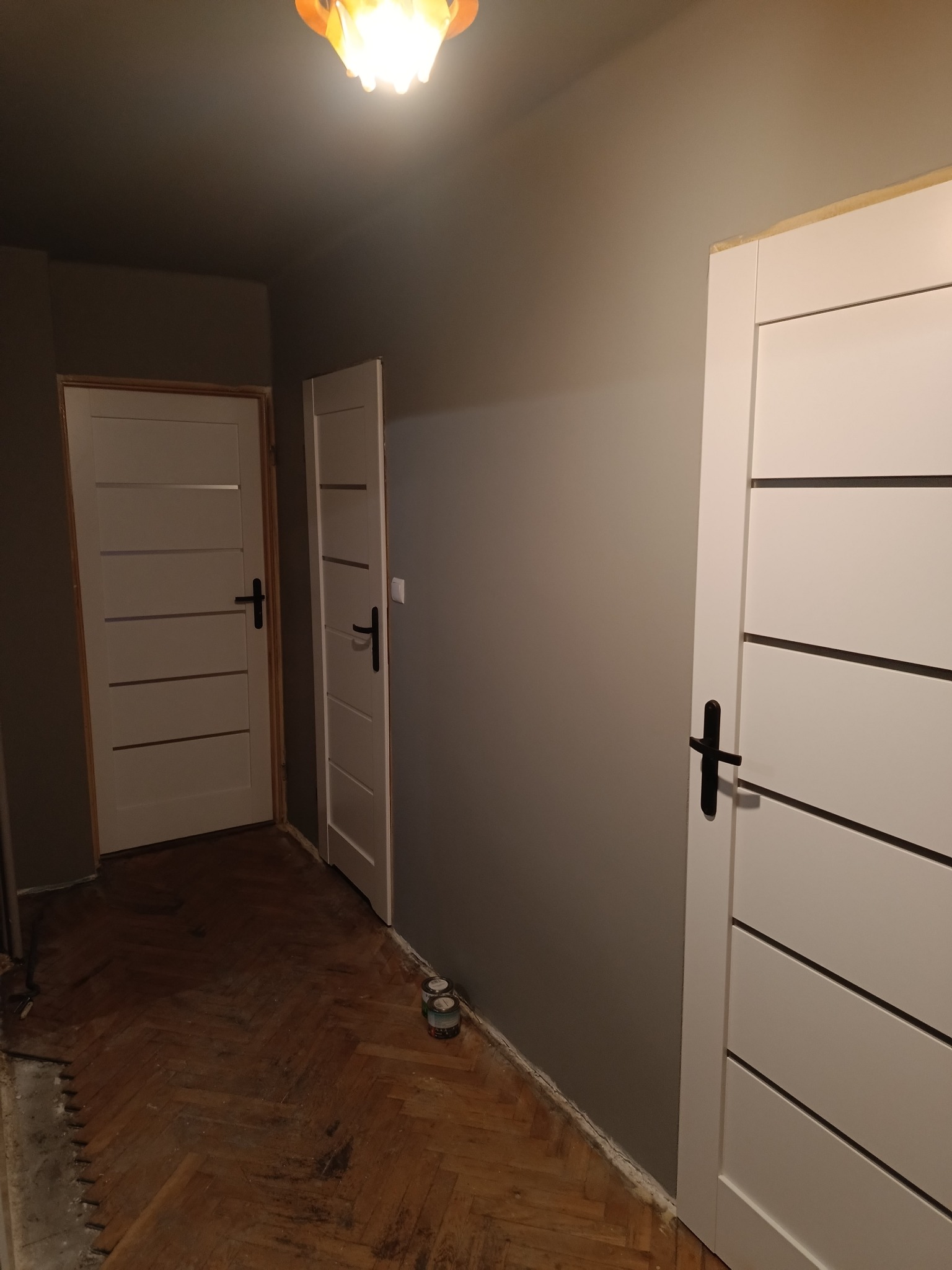 Szare ściany korytarza z dwiema białymi, nowoczesnymi drzwiami z czarnymi klamkami; podłoga w trakcie prac, widoczne puszki z farbą.