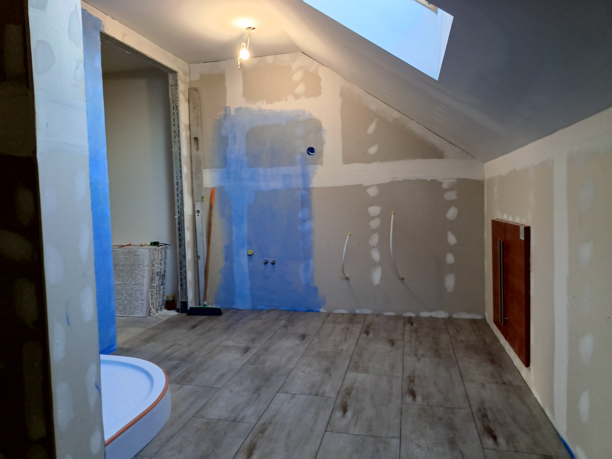 Poddasze w trakcie prac wykończeniowych: ściany z płyt kartonowo-gipsowych, częściowo pokryte gruntem, widoczne instalacje, ułożona podłoga z paneli imitujących drewno, kabina prysznicowa w rogu.