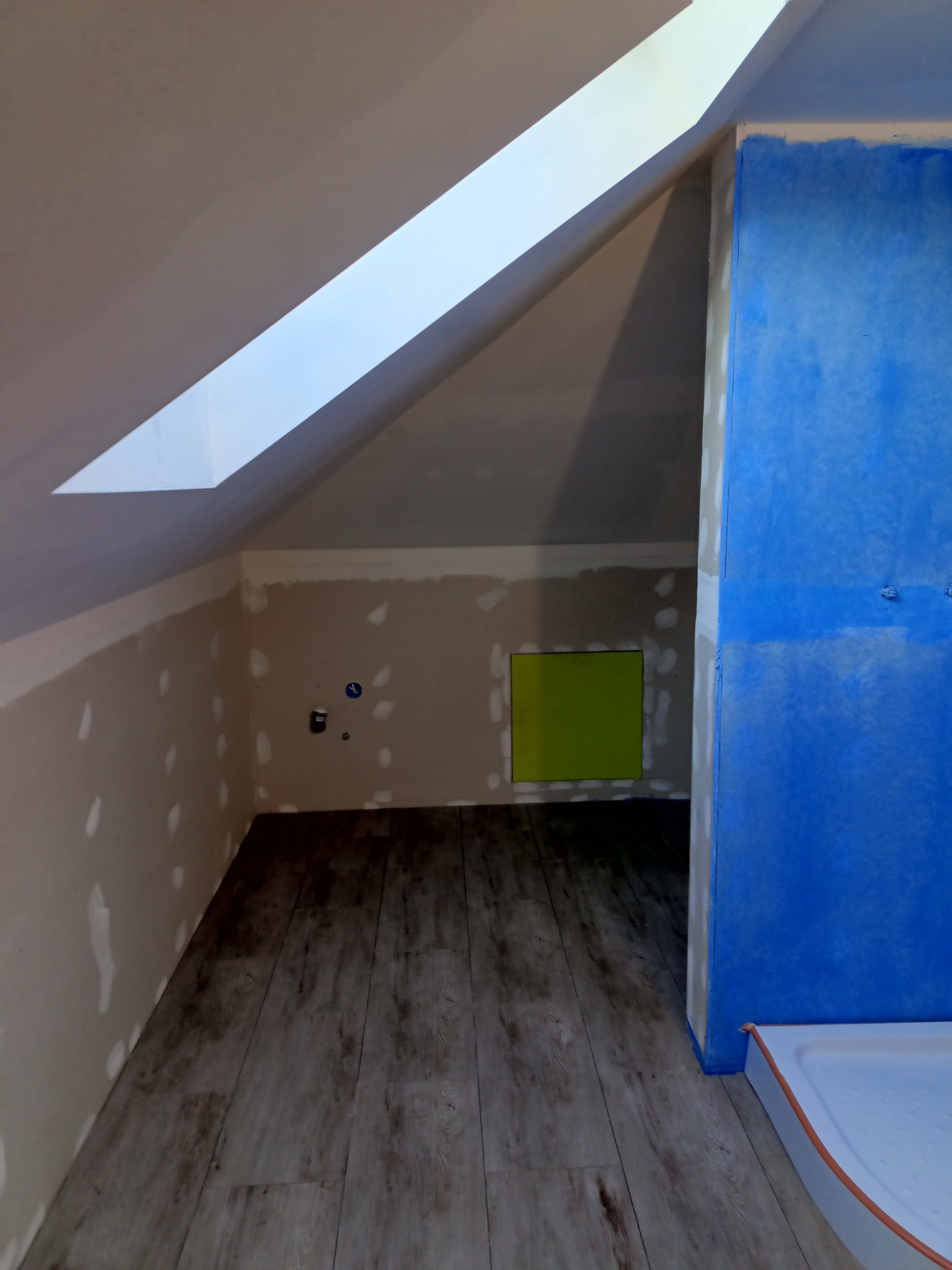 Poddasze w trakcie adaptacji na łazienkę: widoczne płyty karton-gips na ścianach z miejscowymi szpachlowaniami, fragment niebieskiej hydroizolacji oraz ułożone panele podłogowe imitujące drewno.
