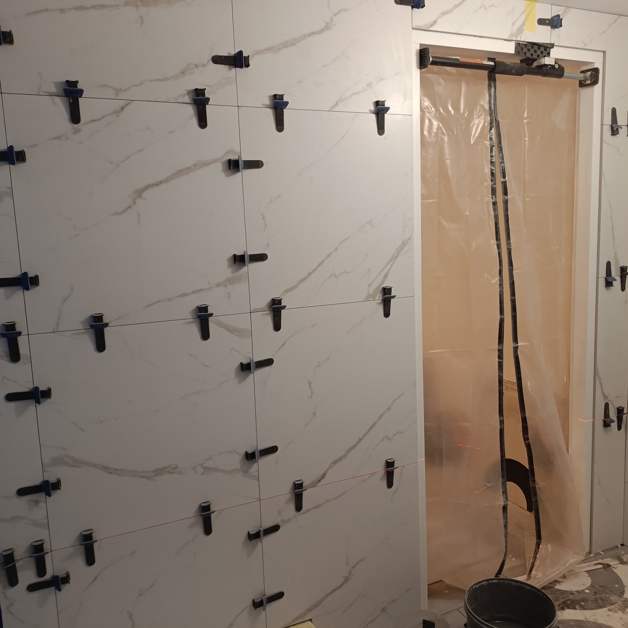 Ściana z białych płytek imitujących marmur, z czarnymi klipsami poziomującymi, widoczna czerwona linia lasera, zabezpieczone folią wejście do pomieszczenia.